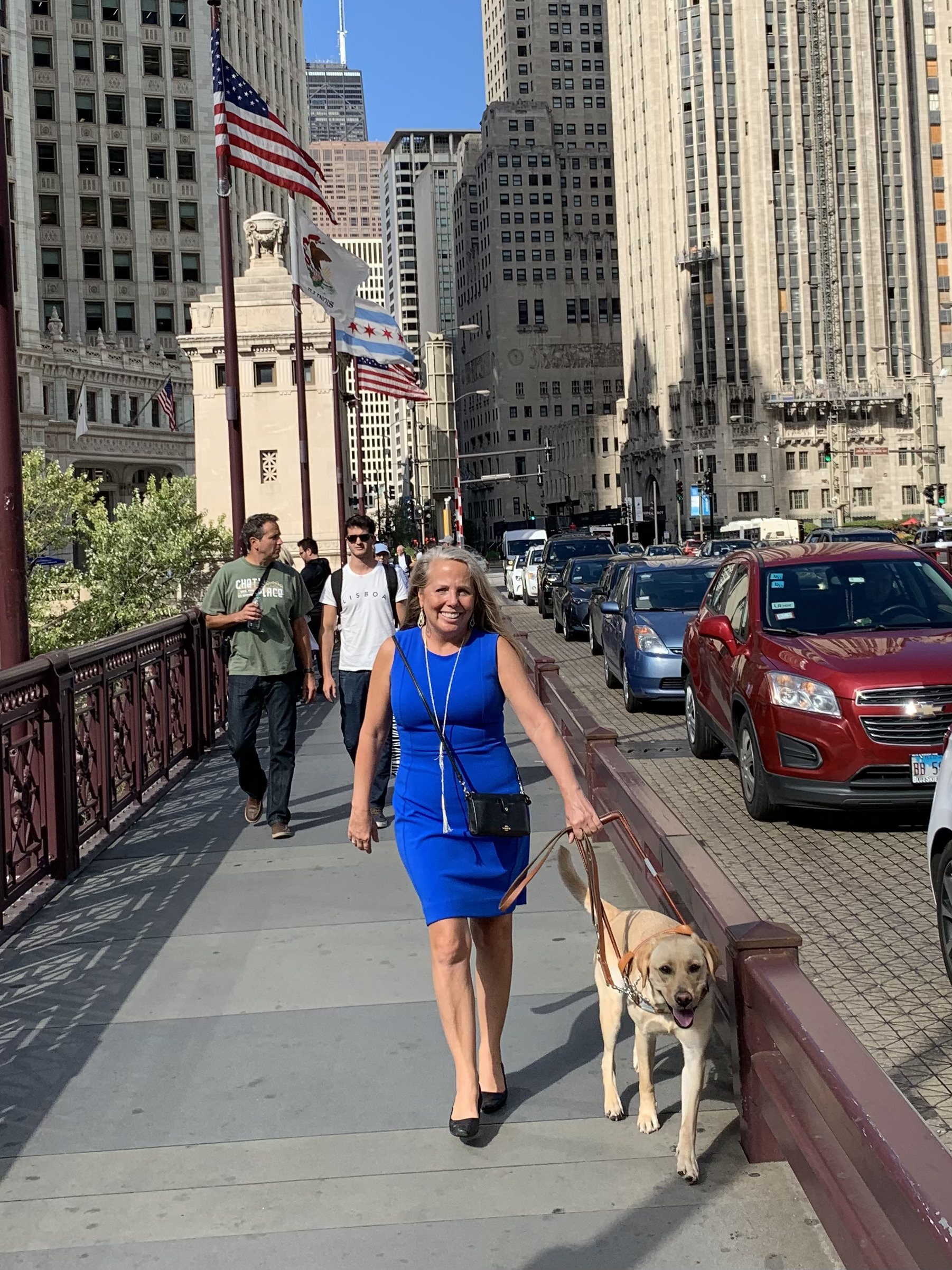 Blonde woman in blue dress with guide dog on busy city street with flags